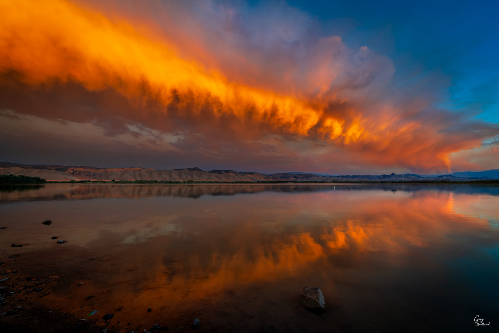 Wildfire smoke illuminated by a vibrant sunset, stretching across the sky and reflecting in calm water beneath distant hills.