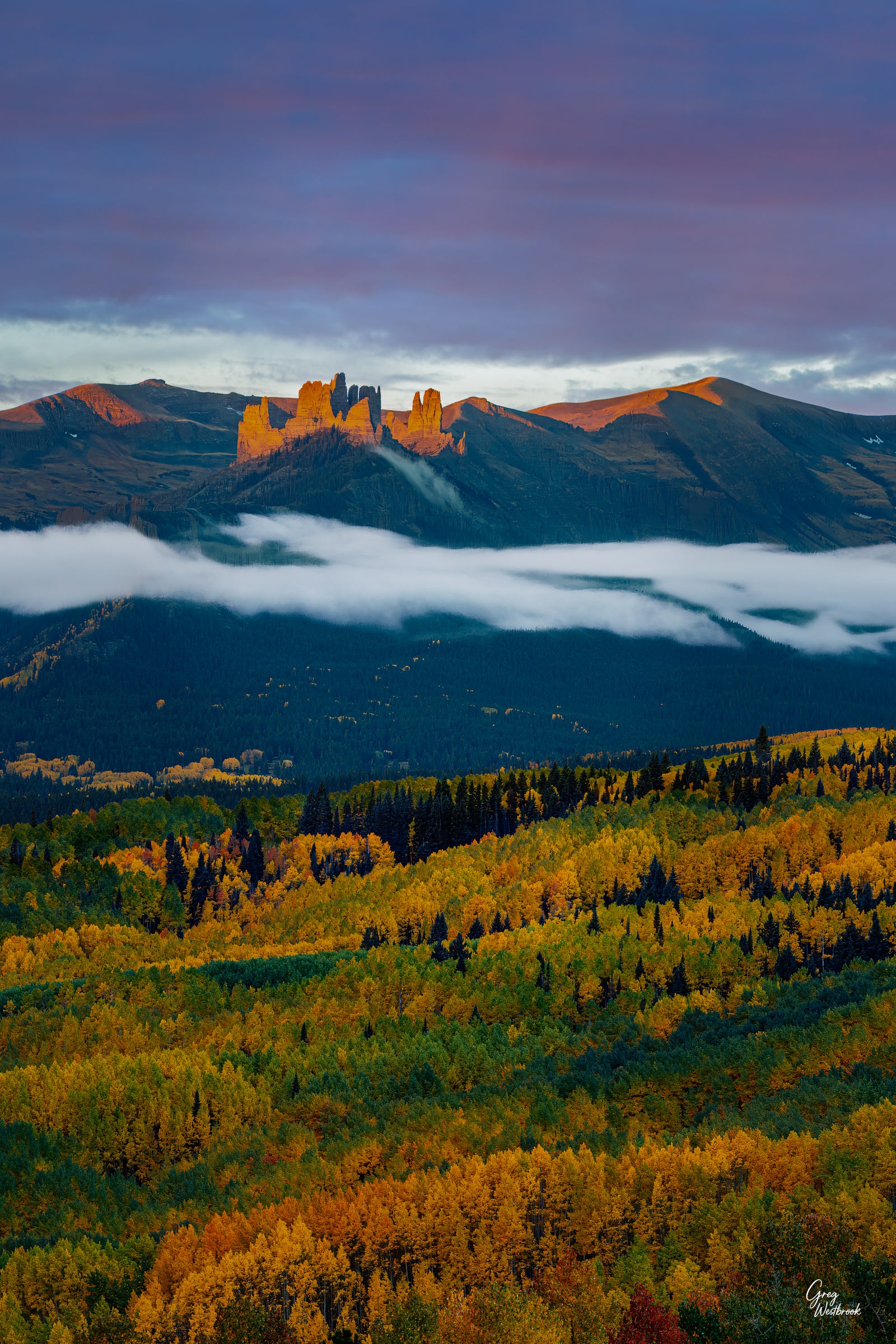 Autumn aspen forest glowing gold beneath low clouds and distant mountain peaks lit by warm sunrise light in a dramatic fall landscape.