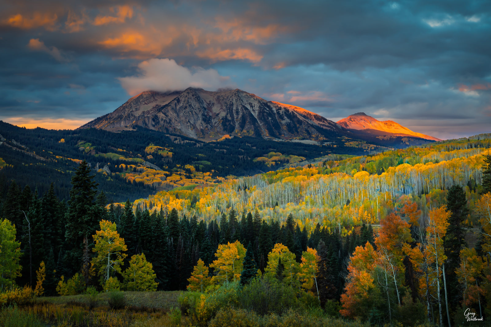 Golden autumn aspens blanket rolling mountain hills beneath dramatic storm clouds as sunrise light ignites the peaks in warm orange tones.